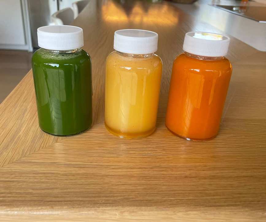 Three small screw-top shot drinking bottles with white lids. One with green liquid, one with yellow liquid, and one with orange liquid.