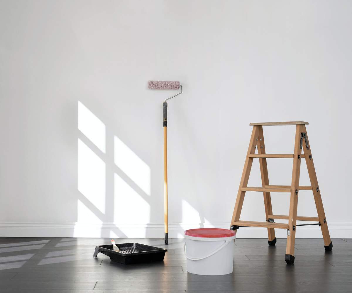A paint roller, step ladder, bucket, and tray on the floor in front of a white painted wall.