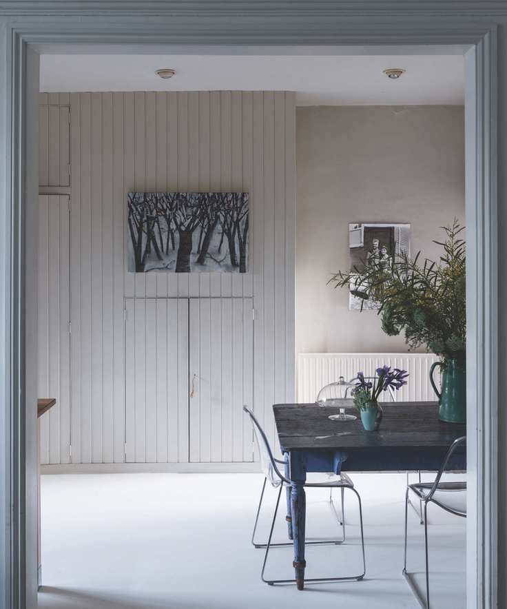 timeless interior design styles, stone and white minimalist kitchen, shiplap cabinetry, vintage table, clear plastic/metal chairs, white floor, huge vase of flowers, artwork