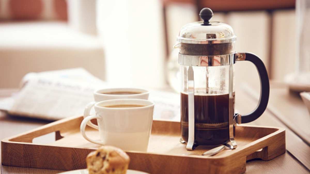 A french press with a muffin and two cups of coffee on a wooden tray
