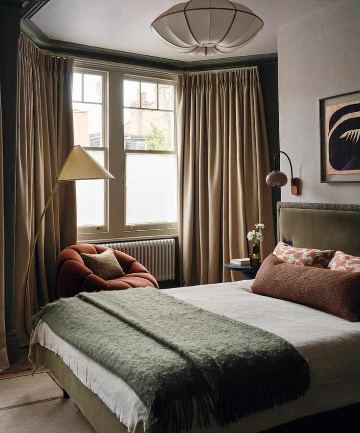 an earthy bedroom with deep green paint in the bay window, neutrals on the rest of the walls, a green velvet bed and a rust red armchair