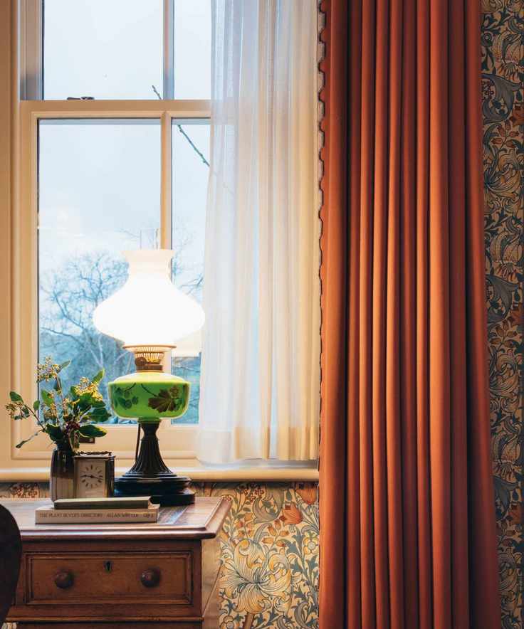 Desk overlooking a window with orange curtains, printed floral vintage wallpaper, wooden desk, antique glass lamp and books