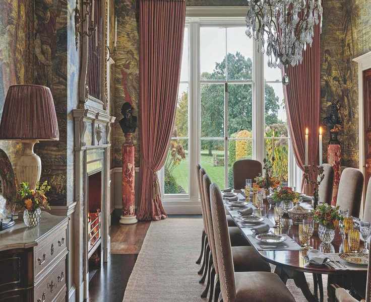 A formal dining room with expansive floor-to-ceiling windows, striking pink curtains, a crystal chandelier, and walls adorned with large classical tapestries