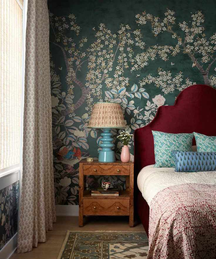 A bedroom with teal floral wallpaper, a burgundy headboard, blue and burgundy bedding, and a wooden nightstand with a blue lamp.