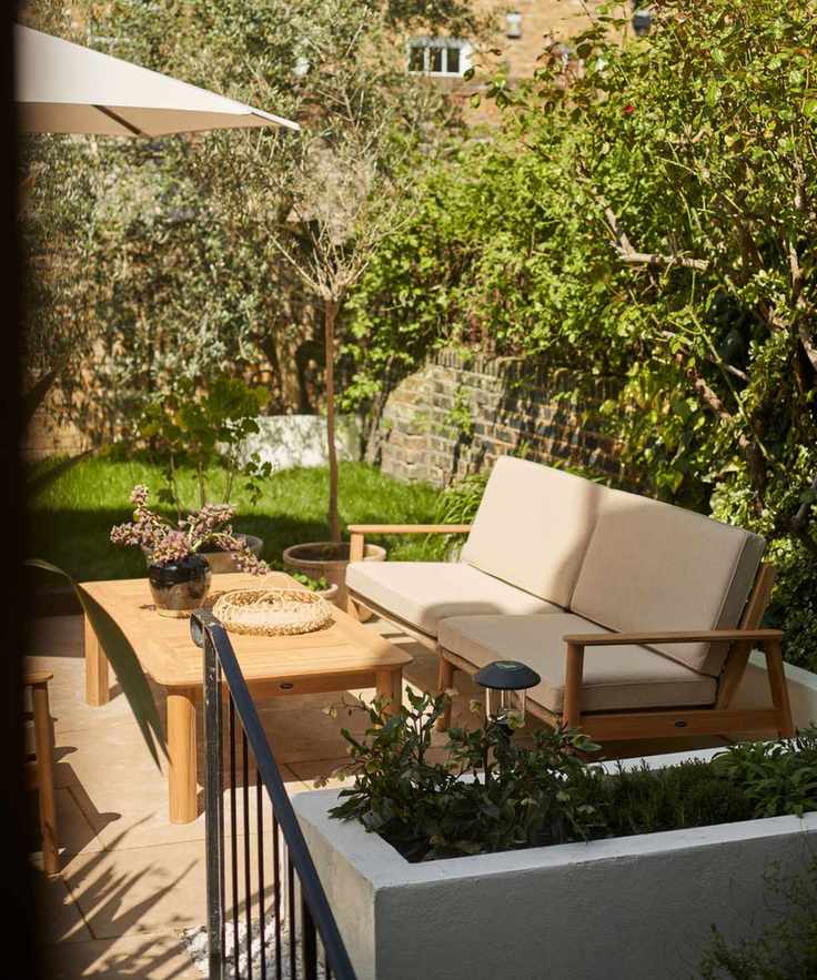 A wooden outdoor sofa with cream upholstered cushions. A wooden outdoor table in front of it, in partial sun.