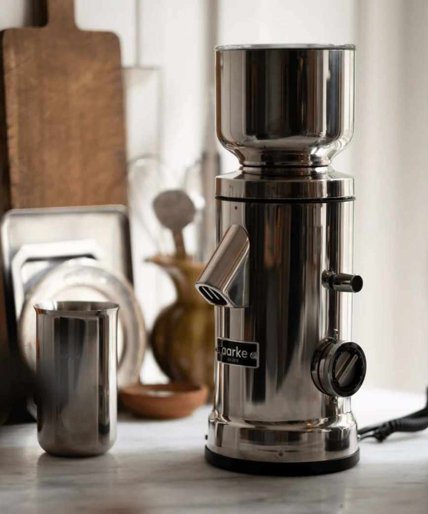 A silver coffee grinder on a kitchen counter beside a stainless steel catch up. A wooden cutting board and baking trays lean behind the wall in the background.