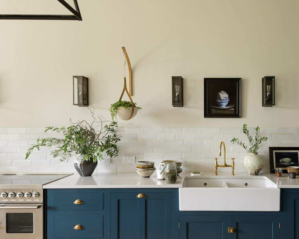 blue and white kitchen, blue cabinetry with white countertops, wall light, counter cabinet with glazing, butler sink, artwork, glass topped stove, brass knobs on cabinets