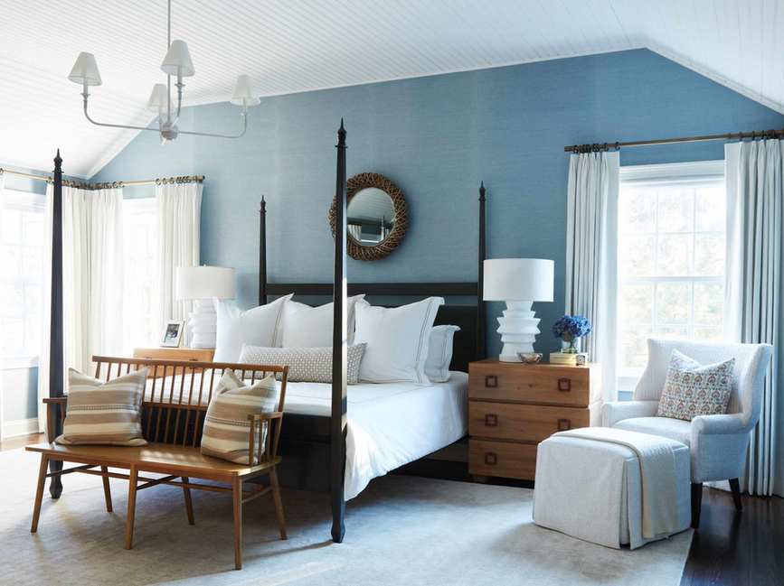 Bedroom with blue grasscloth walls, dark wooden four-poster bed, cream rug, cream armchair and ottoman and wooden bench
