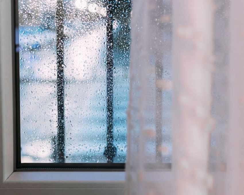 A window with black bars in front of it outside and rain drops on it, with a white thin curtain in front of it inside