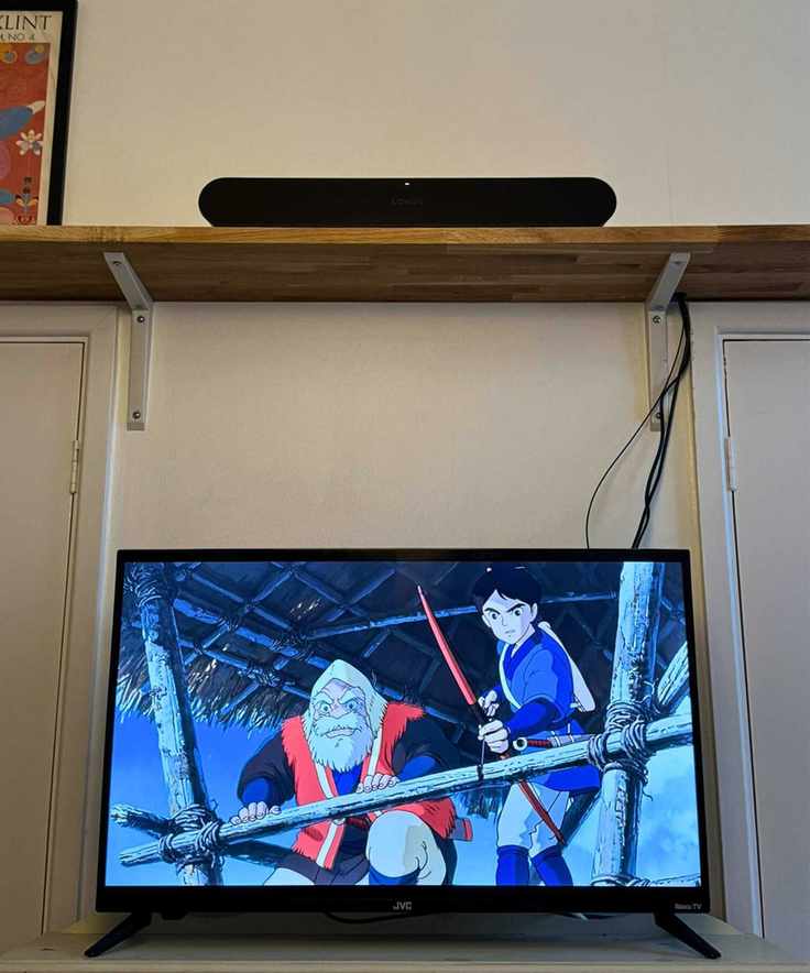 The Sonos Ray soundbar on a wooden shelf above a TV showing Princess Mononoke, in front of cream-white wall.