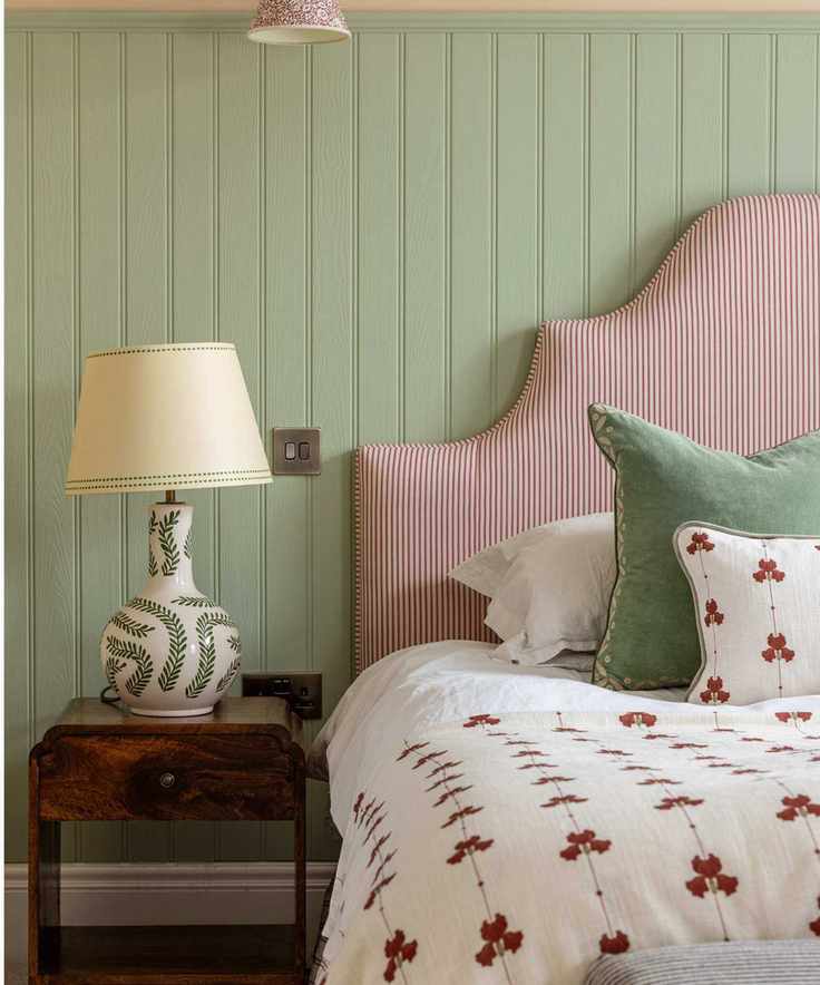 Bedroom with sage green painted wall panelling, a striped upholstered headboard, white bedding and a red and white quilt and throw pillow, a mahogany nightstand with a green and white lamp on top