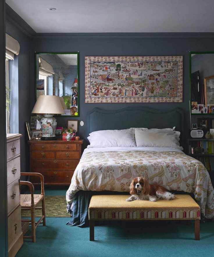 Cozy bedroom with a dog resting on a footstool. A beautiful landscape painting hangs above a bed adorned with patterned sheets, next to a lamp and an armchair.