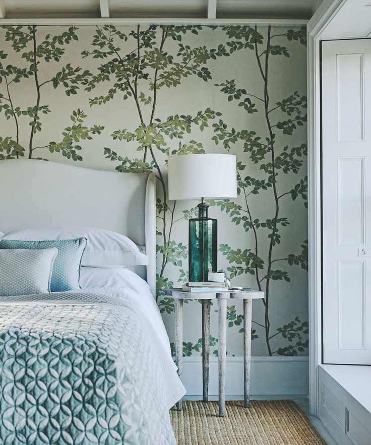 A wallpapered bedroom with green trees on it, a large white bed with blue bedding, wooden bedside table with green blue glass lamp with white lampshade, and large white shutters seen to the right. The carpet is textured and woven.