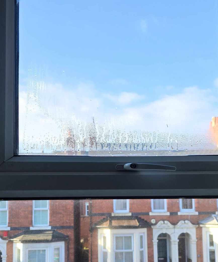 A close up of a window overlooking a blue sky with clouds and a row of brick houses with white windows and dark colored doors. There are lots of droplets of condensation on the lower 2-3 inches of the glass