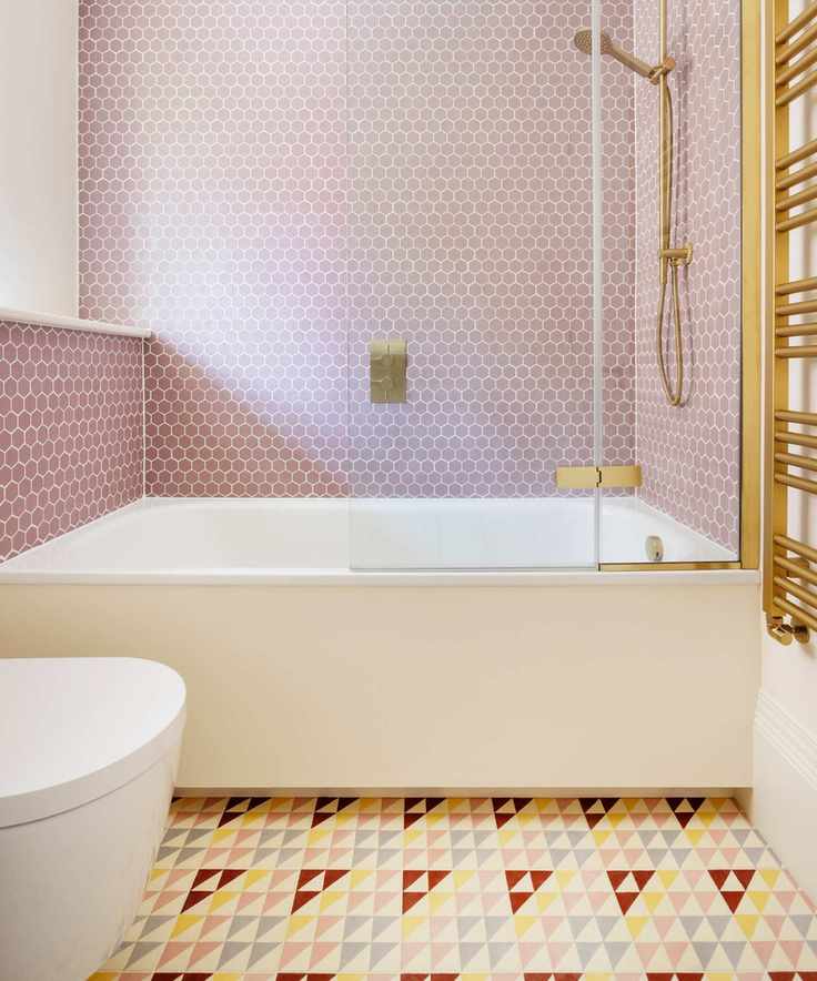 small pastel bathroom with small pale pink hexagonal wall tiles, brighter yellow, blue and red floor tiles, brass towel warmer, brass shower fitting, small bath, shelf