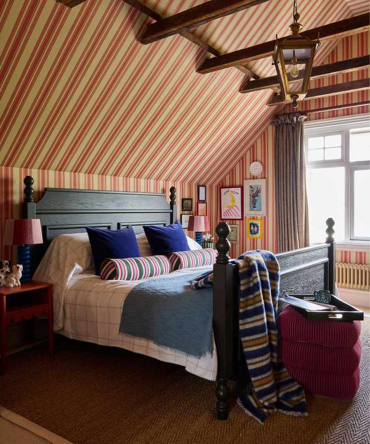bedroom with a sloped ceiling and beige and red striped wallpaper that goes up and over the ceiling. A large dark wooden vintage bed sits in the middle styled with striped throws and pillows with a velvet stool at the foot of the bed