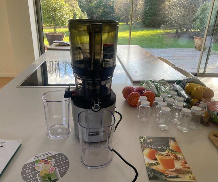 A silver based cold-press juicer with a grey plastic top, set up with two drinking glasses to the front and side. An instruction manual beside it.