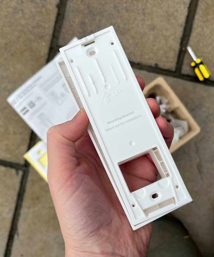 Hand holding the back plate of the Tapo D210 Video Doorbell above other parts and tiled concrete floor.