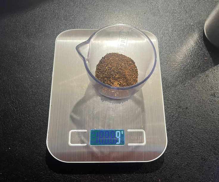 Coarse ground coffee beans in a clear plastic container on digital weight scales.