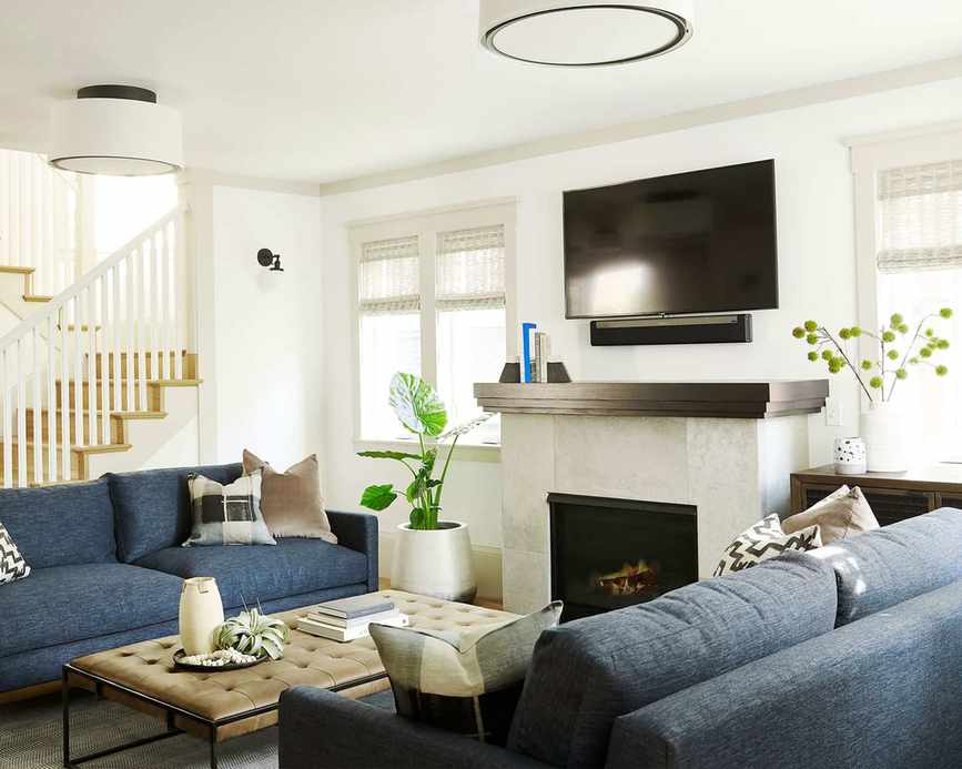 Bright living room painted white features a stone fireplace with a TV mounted above, twin blue sofas, and a central low leather coffee table adorned with plants and cushions.