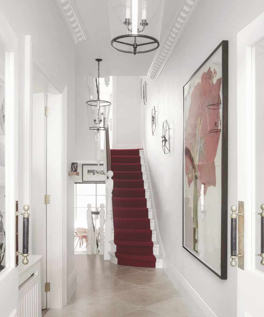 A white entryway with a white staircase and red stair runner, framed artwork, and glass lamps.