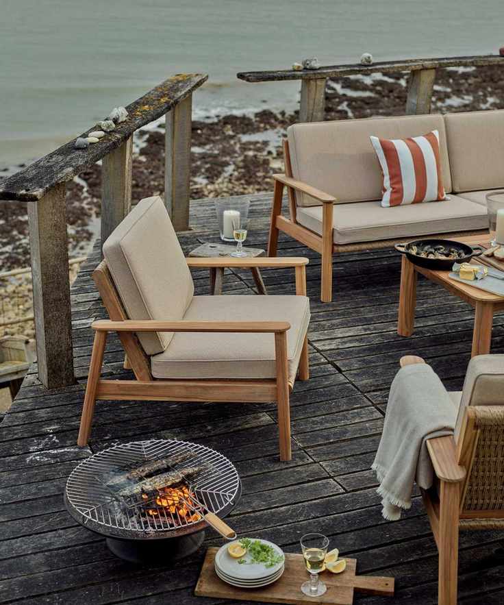 A wooden outdoor armchair with cream upholstered cushions on a wooden deck beside the sea. A small BBQ firepit beside it.
