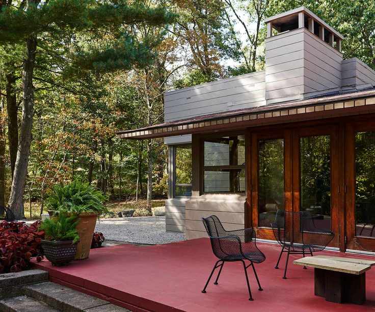 Midcentury house with red patio, iron chairs and wood table and trees surrounding the grounds