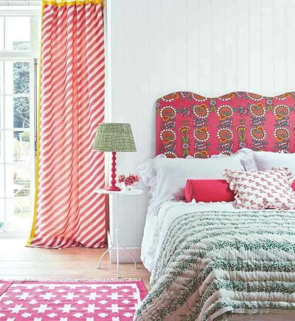 A colorful bedroom with white panelled walls, wood floors, pink patterned headboard, cushions and rug, white bedding, and green bedspread. Large window with red and white striped curtains on the left.