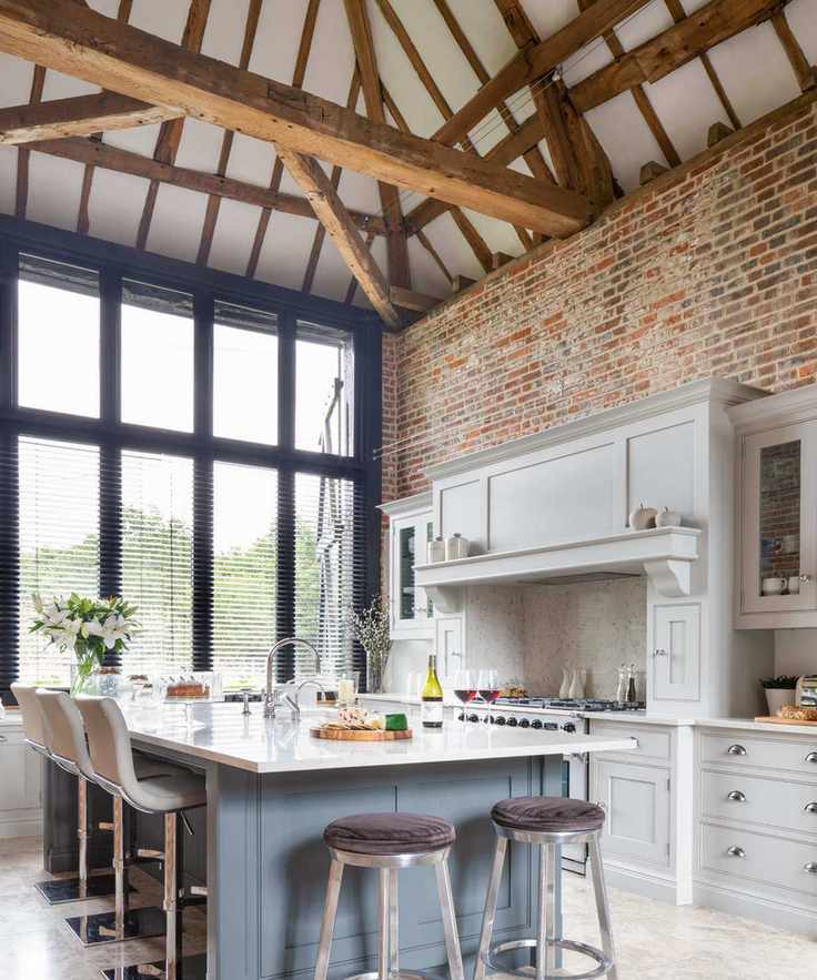 Open plan kitchen in a tall beamed space with exposed brick wall, pale gray cabinetry and a kitchen island.