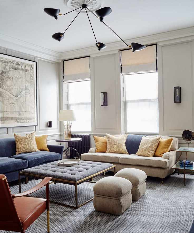 Living room with pale gray walls, cream and blue sofas, a dark gray fabric ottoman with a metal frame, and two gray poufs beside it.