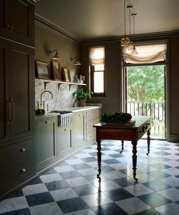 A moody kitchen with dark green cabinets, checkerboard floor tiles, marble countertops and backsplash, and a traditional work table island with brass castors