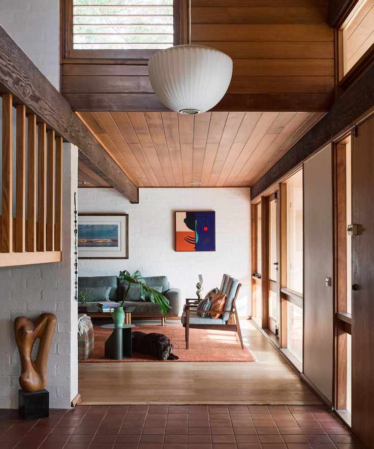 a hallway leading into a living room with mid-century furniture, art, large windows to the right, wood floors and ceiling, and a dog on the floor