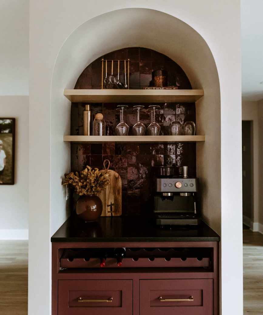 A small home bar installed into an arched alcove with wine red glossy tiles and cabinetry paired with brass shelving