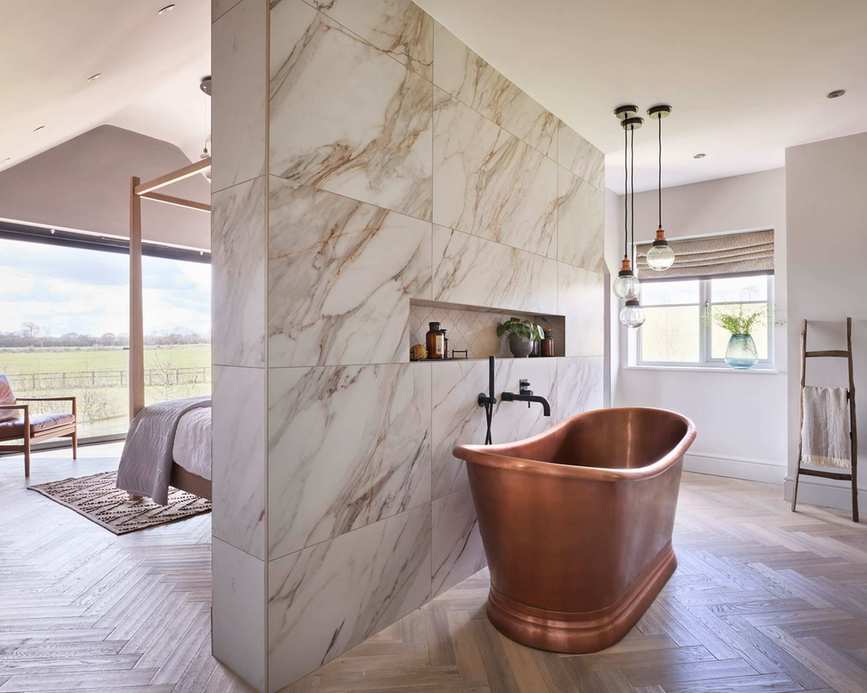 Large, modern bedroom with bathtub, separated by marble partition wall, copper bathtub, wooden flooring, trio of hanging pendant lights, bed and leather and wooden armchair in background, large floor to ceiling windows
