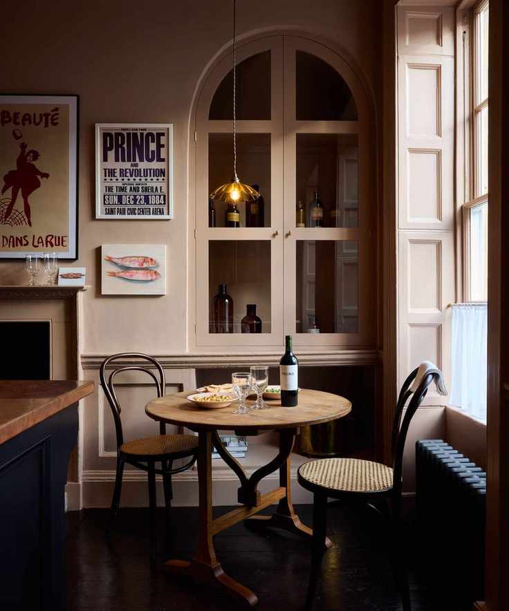 French bistro style kitchen with small round dining table, wooden bistro chairs, pink wall with cabinet and low pendant light