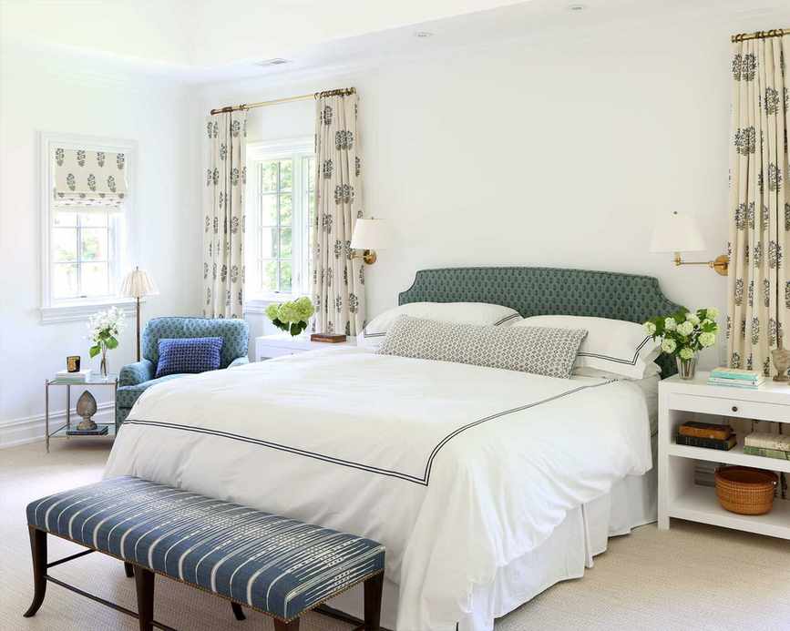 Master bedroom with patterned curtains and matching blind, large bed with white bedding, blue patterned headboard, bench at end of bed, cream carpet, white painted walls, two bedside tables, chair in corner with matching upholstery to headboard