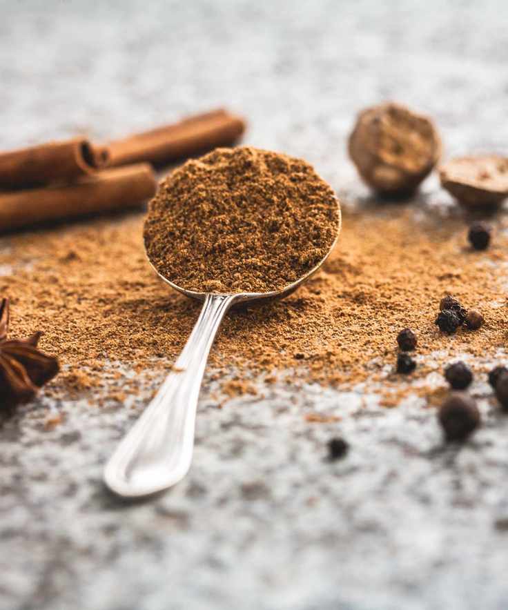 Silver spoon of cinnamon powder, on a granite surface, with loose cinnamon powder and sticks in view