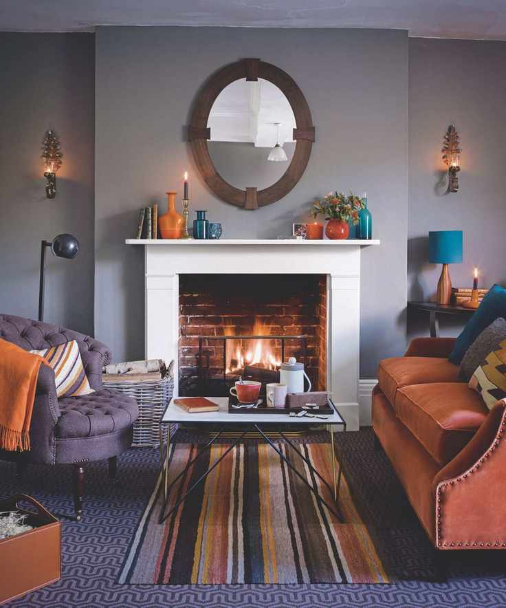 A lilac living room with a white mantelpiece, orange and purple chairs and sofas with matching cushions, and a striped rug underneath a coffee table in the center of the room.