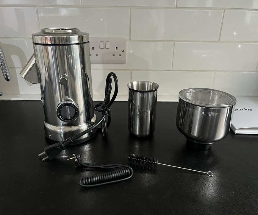 The parts of the Aarke coffee grinder, including the mechanical base, stainless steel grounds cup, shopper with glass lid, cleaning brush, and connector cable, on a black counter.