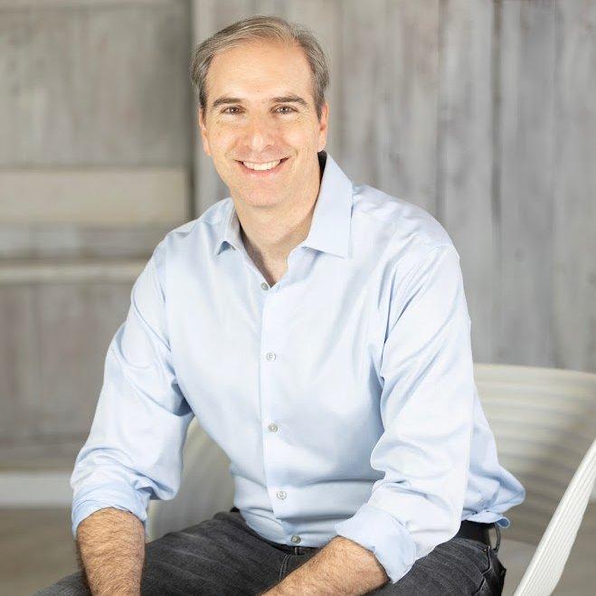 a photo of professional organizer Ben Soreff sat on a white chair against a wooden background, wearing a light blue collared long sleeve shirt and black jeans