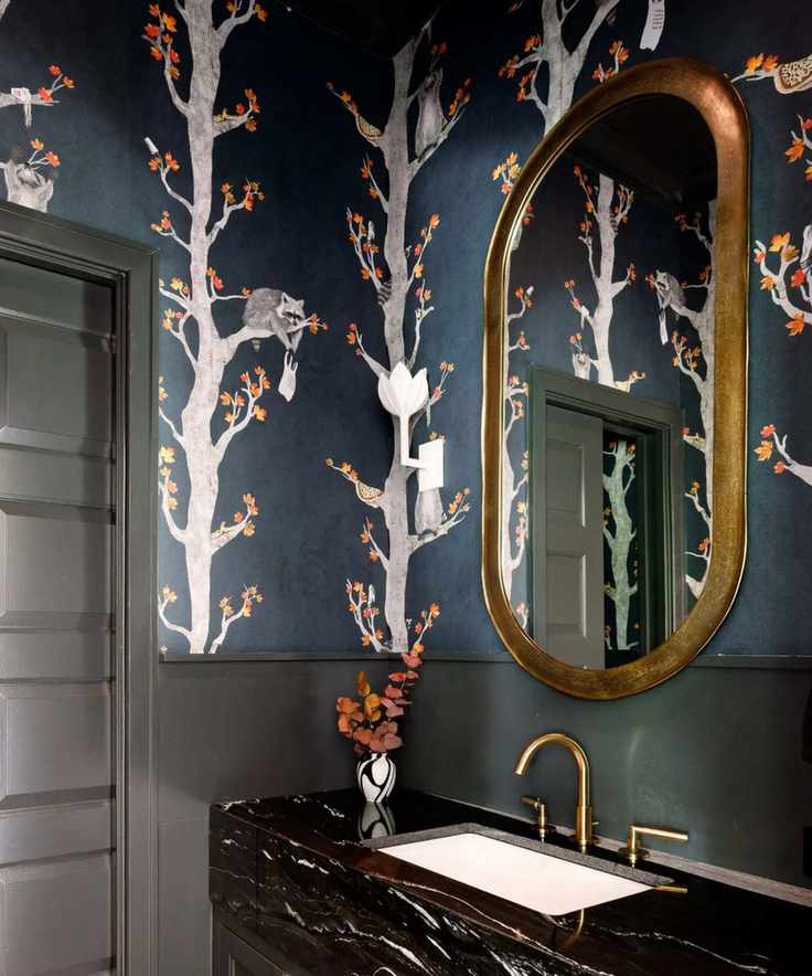 A small bathroom with a black marble sink on a black vanity and moody blue wallpaper depicting trees