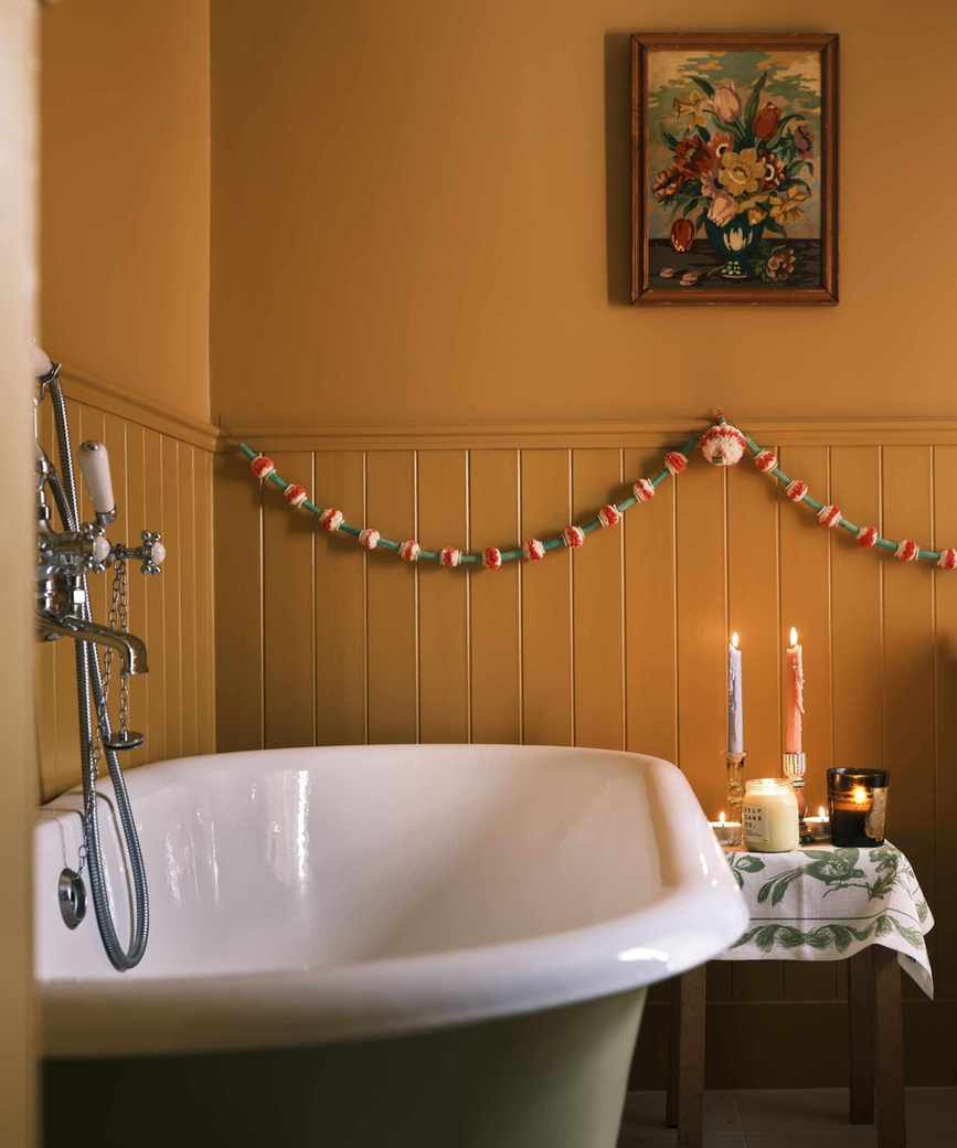 Mustard yellow bathroom with cozy candles and festive bunting. The roll top tub is sage green on the outside, the walls are panelled on the bottom half and there is a framed painting of a vase with flowers on the wall. It feels festive and cozy