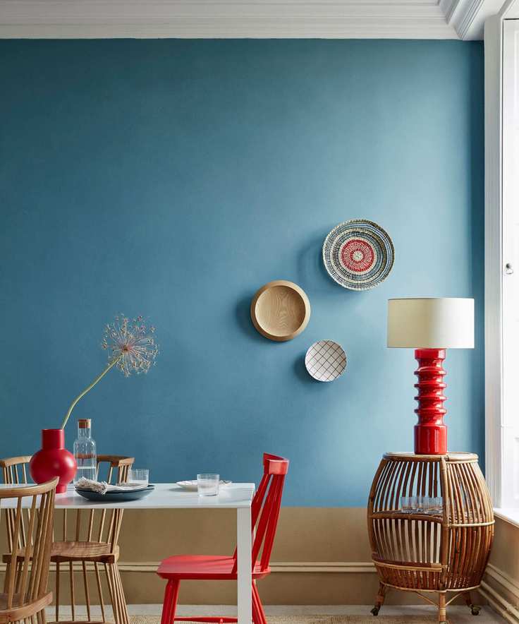 A blue walled dining room with red dining chairs and red based floor lamp. Three baskets hung on the wall