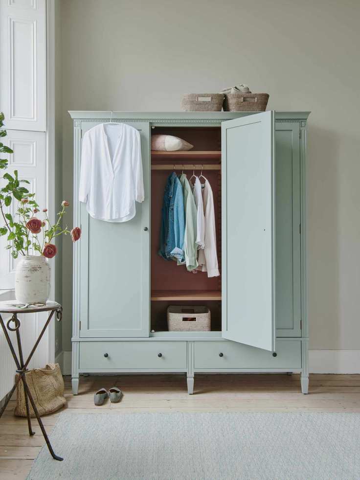 bedroom with aqua freestanding wardrobe, clothes hanging, baskets on top, side table near window with flowers, aqua rug