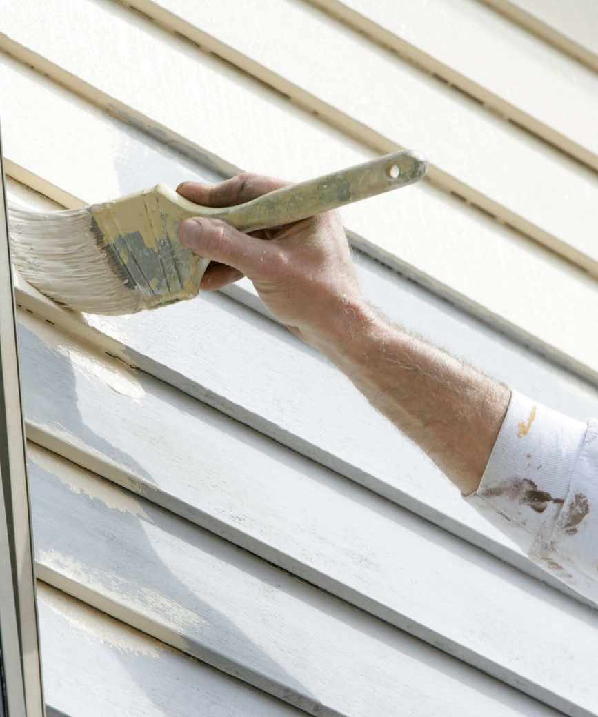 A mans hand painting wooden siding on a house white with a paintbrush.