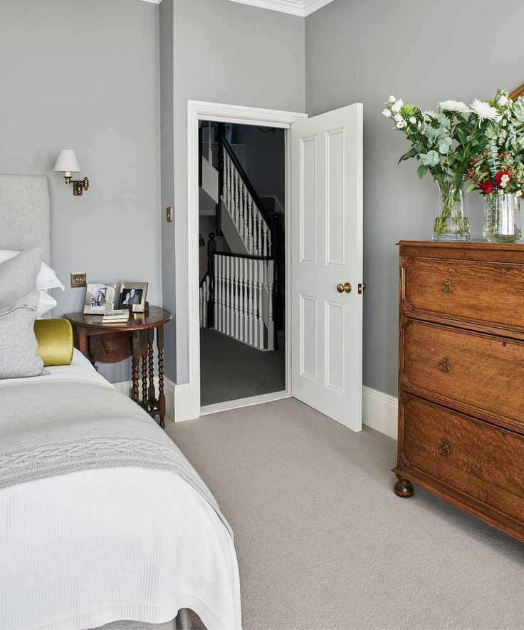 A grey bedroom with a double bed with white bedding seen to the left, a brown bedside table with framed photographs, grey walls and a large brown wooden dresser with glass vases of flowers. The bedroom door is open to show a black and white balustrade behind.