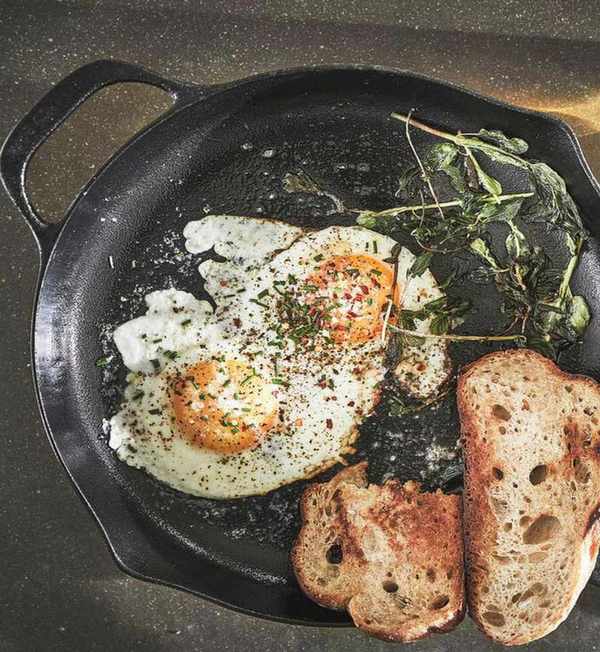 A black cast iron skillet on a black worktop, with two fried eggs, two slices of rustic toast, and some wilted spinach inside.