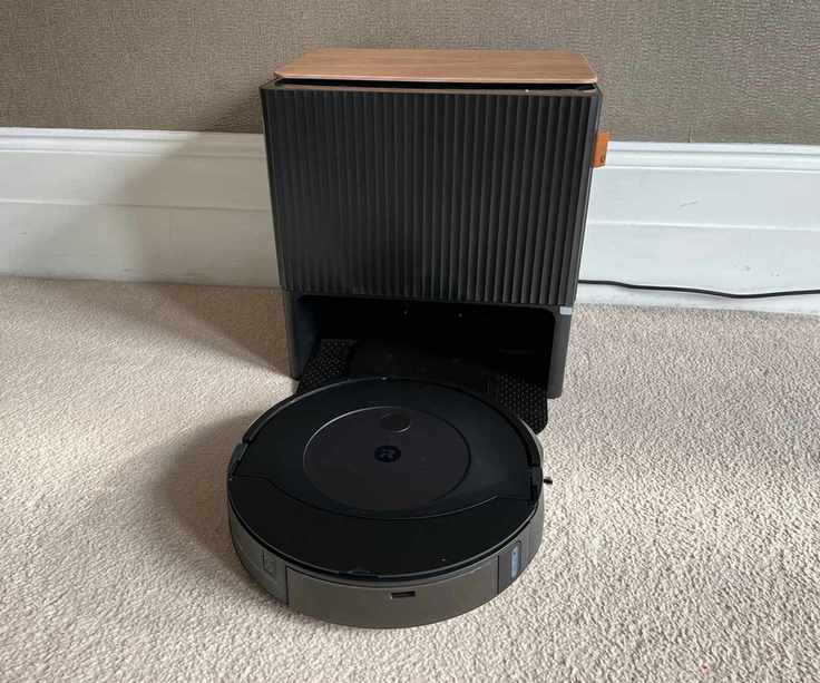 The iRobot Roomba Combo j9+ Robot Vacuum and clean base on cream carpet in front of white baseboard and brown wall in Alex's apartment, with power cord going off-image to the right