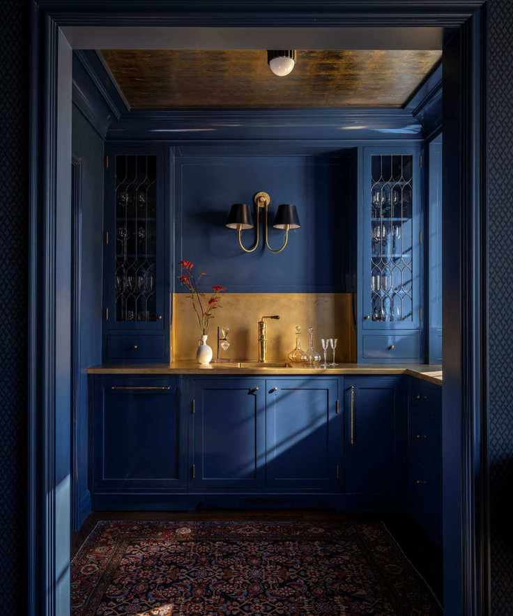 Bar pantry with dark blue cabinetry and walls, gold accents, glassware, and a red Persian rug
