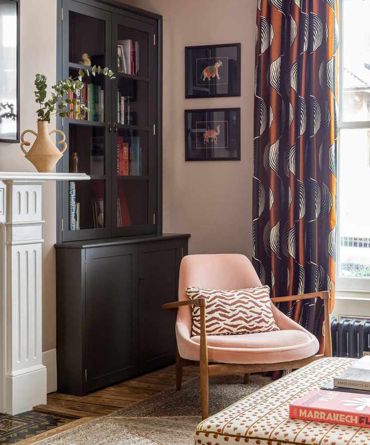 Eclectic living room with woven rug, pink wood armchair, ottoman, bookshelf, orange printed curtains and wall art